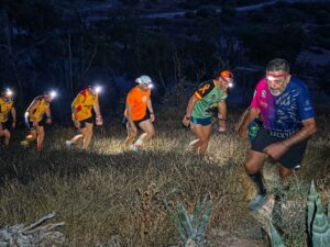 Group of night trail runners with headlamps climbing a hillside; rugged, dry vegetation around them.