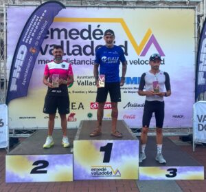 Three male winners stand on a podium holding plaques: 1st place center, 2nd on the left, 3rd on the right, with a colorful event backdrop behind them.