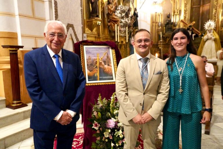 Three people in formal attire stand inside a richly decorated church beside a framed poster on a red-draped stand and a bouquet of flowers.
