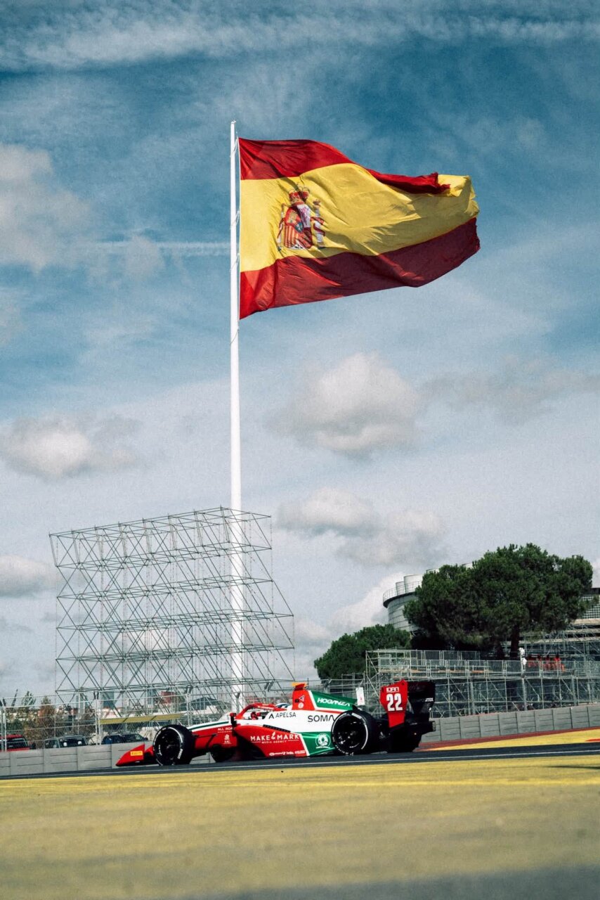 Large Spanish flag waves atop a tall flagpole at a racing circuit, with a red-and-green Formula 1 car on the track in the foreground.