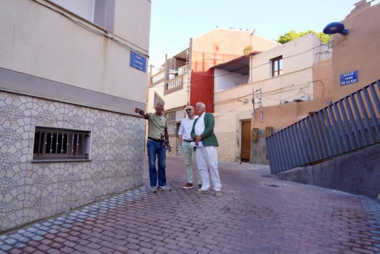 Three men stand in a narrow cobblestone street, one pointing toward the left as the others look on, with pastel buildings and a blue railing in view behind them.