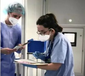 Two healthcare workers in scrubs and masks review documents on a clipboard in a clinical hallway.