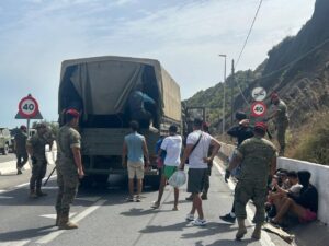 Soldiers in camouflage with red berets surround a loaded military truck stopped on a road as civilians wait nearby under a 40 km/h sign on a sunny day.