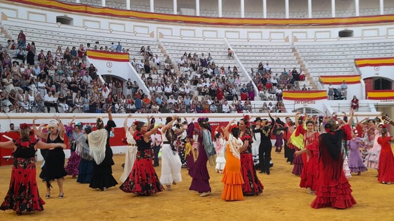 Group of flamenco dancers in colorful dresses performing in a circular arena with a seated audience nearby.