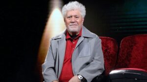 An older man with white hair and a light gray beard sits in red theater seats on a stage, wearing a gray jacket over a red polo shirt, facing the camera.