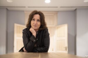 Woman in a black leather jacket leans on a wooden table, chin resting on her hand, in a softly lit room.