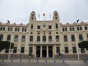 Front view of a beige neoclassical government building with two towers, arched windows, and flags atop, reached by stairs and bollards.