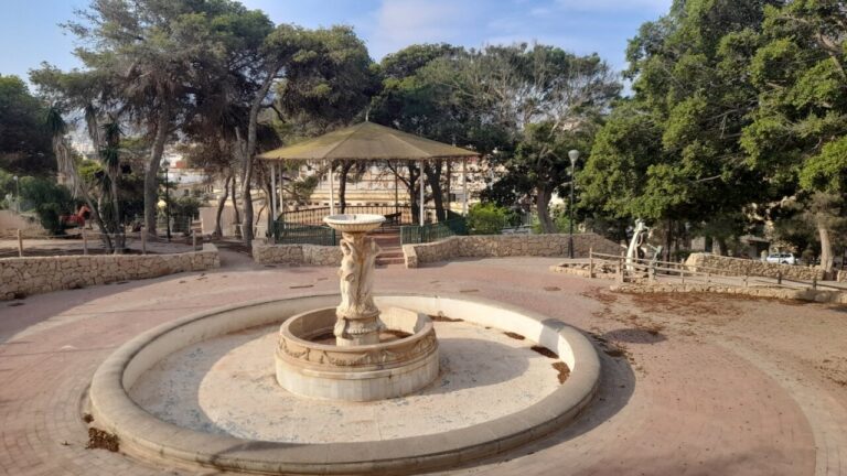 Round stone fountain centerpiece in a park, with a banded circular basin and a sculpted pedestal under a small canopy gazebo nearby.