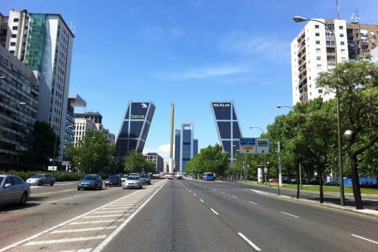Wide city avenue in Madrid with two leaning bank towers labeled Bankia and Realia, cars and buses on the road, green trees, and a clear blue sky.