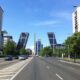 Wide city avenue in Madrid with two leaning bank towers labeled Bankia and Realia, cars and buses on the road, green trees, and a clear blue sky.