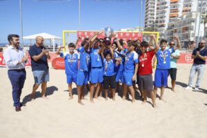 Youth soccer team on a sandy beach celebrating with a trophy and medals in front of a goal, with cheering adults nearby under sunny skies.