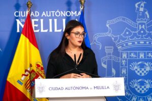 Woman in black shirt and glasses speaks at a podium in front of a Melilla city backdrop with flags on either side.