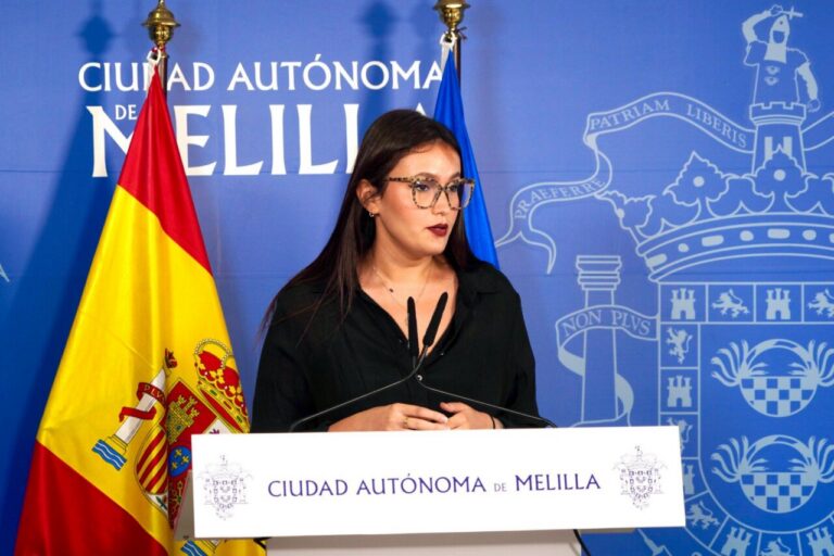 Woman in black shirt and glasses speaks at a podium in front of a Melilla city backdrop with flags on either side.