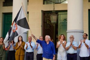 El presidente Juan Vivas ondeando la bandera de Ceuta con un grupo de personas aplaudiendo.