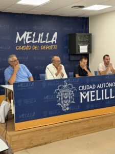 Four men sit at a panel table during a Melilla press conference, blue backdrop reads 'MELILLA' and 'CIUDAD DEL DEPORTE'.