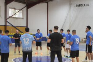 Indoor handball team huddles in a gym, players in blue shirts forming a circle around a coach.