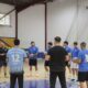 Indoor handball team huddles in a gym, players in blue shirts forming a circle around a coach.