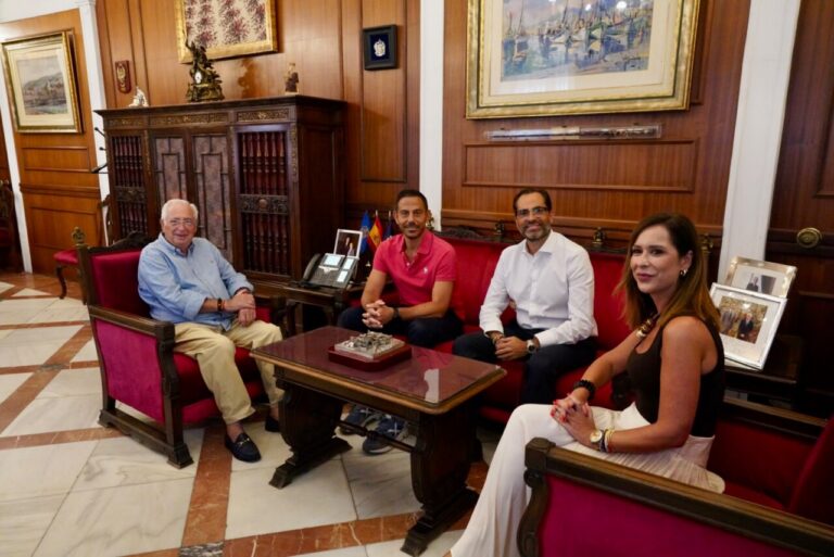 Four adults sit on red upholstered seating in a wood-paneled office around a glass coffee table, posing for a meeting photo.
