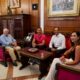 Four adults sit on red upholstered seating in a wood-paneled office around a glass coffee table, posing for a meeting photo.