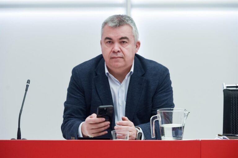 Man in a navy blazer sits at a conference desk, holding a smartphone, with a glass pitcher of water nearby.
