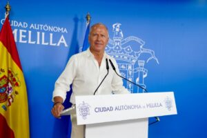 Man in a white shirt speaks at a podium during a Melilla government press conference, with a blue backdrop and the Spanish flag nearby.