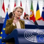 Nueva normativa de la UE sobre pagos en efectivo - MelillaHoy Older woman in an olive blazer speaks at an EU Parliament podium, microphones in front, flags of the EU and member states behind her.