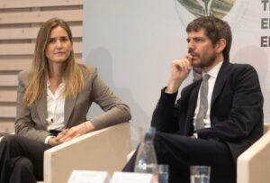 Two professionals in a panel discussion: a woman in a gray blazer and a man in a dark suit seated on stage, microphones out of view.