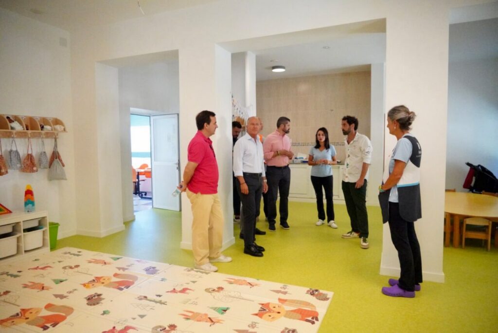 Group of adults standing in a bright daycare playroom, discussing near a colorful animal-themed mat.