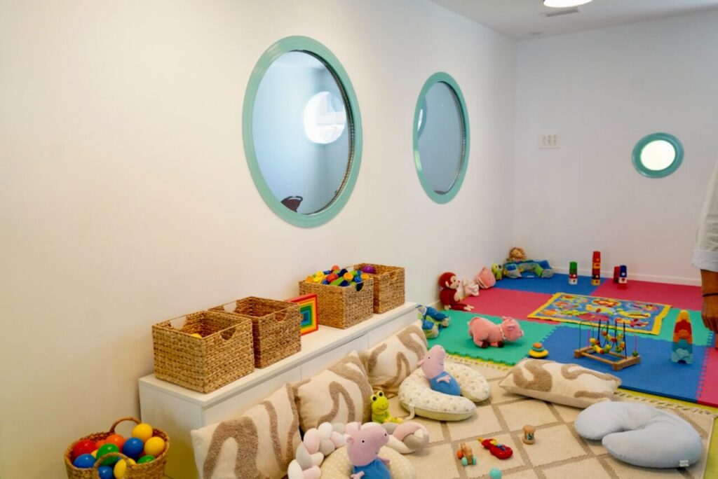 Children's playroom with colorful toys, stuffed animals, and woven baskets on a white bench; round turquoise mirrors on the wall and a foam mat area for play.