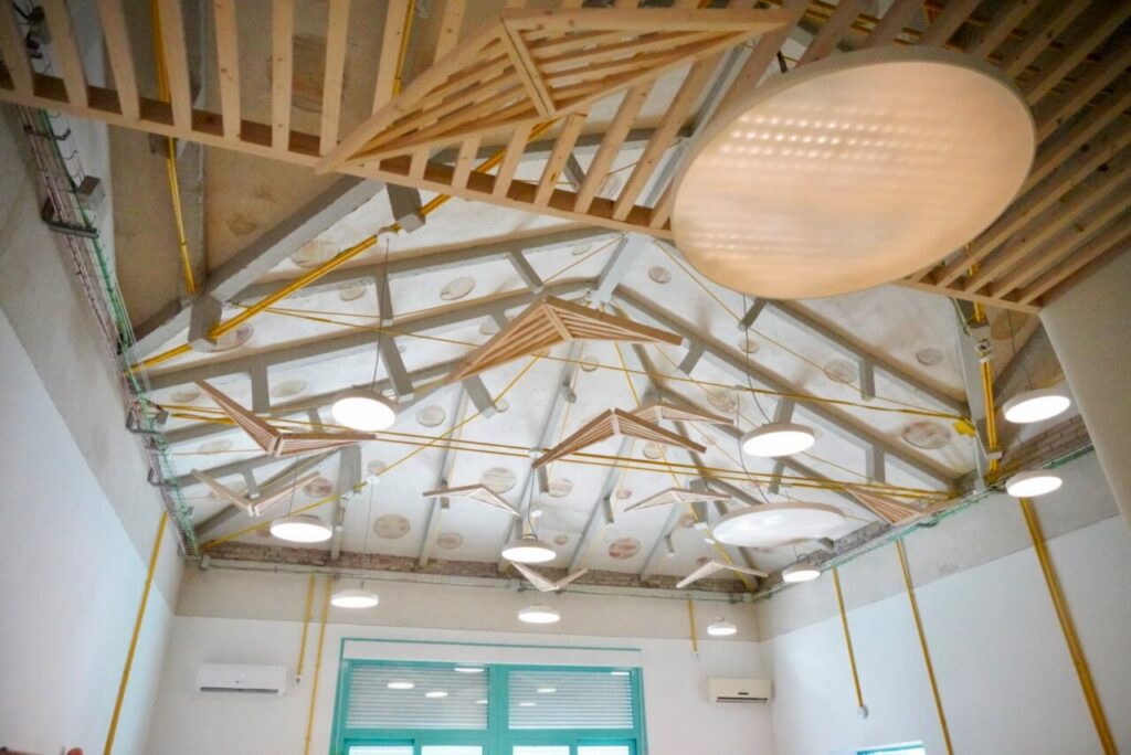 Interior ceiling view with exposed wooden trusses and suspended geometric wooden elements, illuminated by large round pendant lights in a bright room