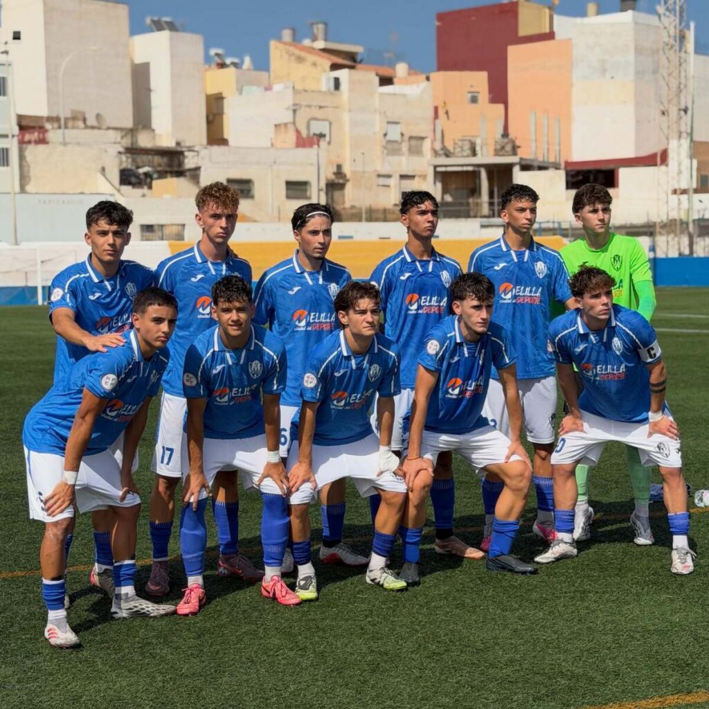 Youth soccer team in blue uniforms posing for a group photo on a turf field, goalkeeper in green on the right, colorful buildings in the background