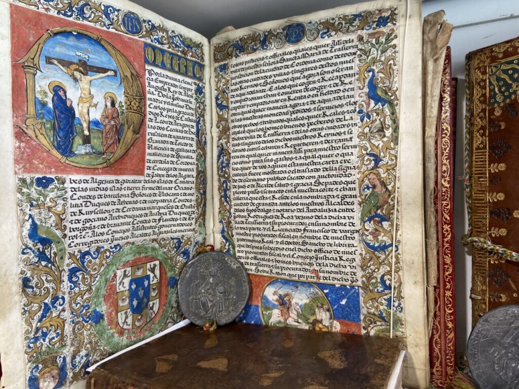 Open illuminated manuscript with ornate gold borders; crucifix scene on the left page and dense text on the right, coin resting on the page edge.