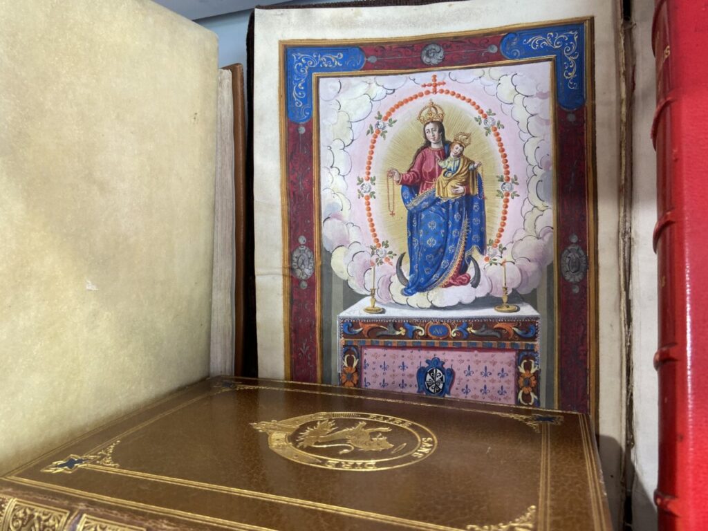 Devotional print of the Virgin Mary holding baby Jesus, framed with ornate blue and gold decoration, resting on a shelf with other books.