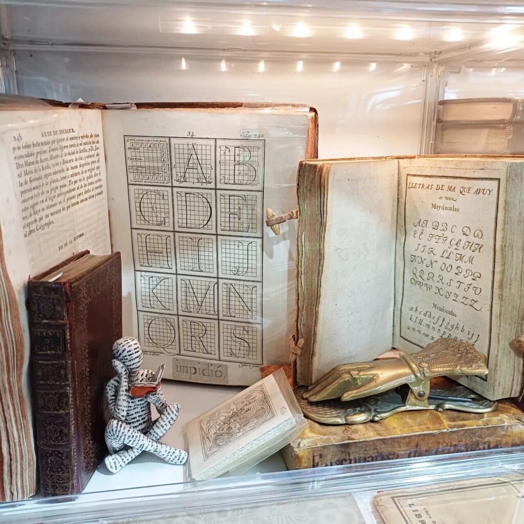 Vintage bookshelf with old books, an alphabet chart, and a small paper-ink figure reading a book; a carved metal object lies nearby.