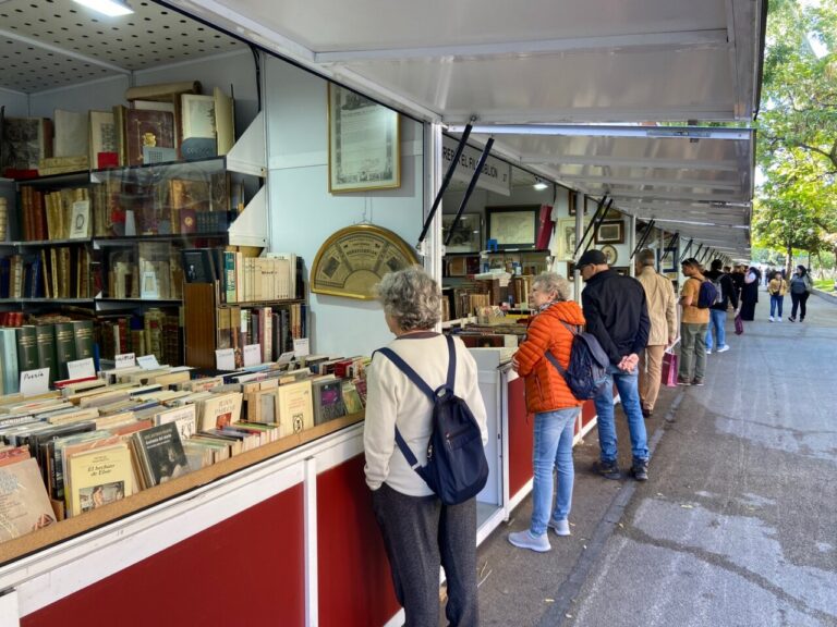 People browsing a long row of outdoor book stalls under white canopies, shelves filled with books and vintage items.