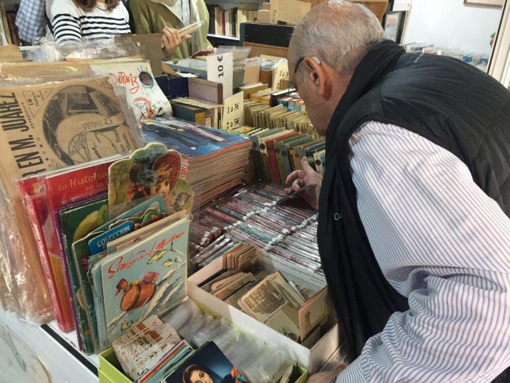 Older man browsing a cluttered book/record shop, sorting through magazines and comics at a display shelf while other customers look on in the background
