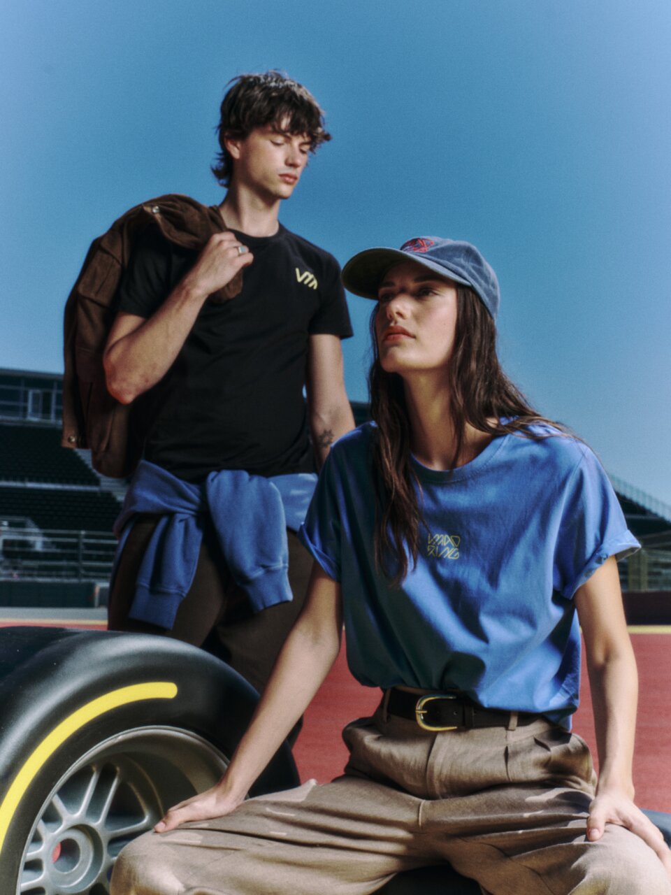 Two young adults at a race track: a woman in a blue shirt and cap sits beside a large tire while a man stands behind her with a jacket over his shoulder.