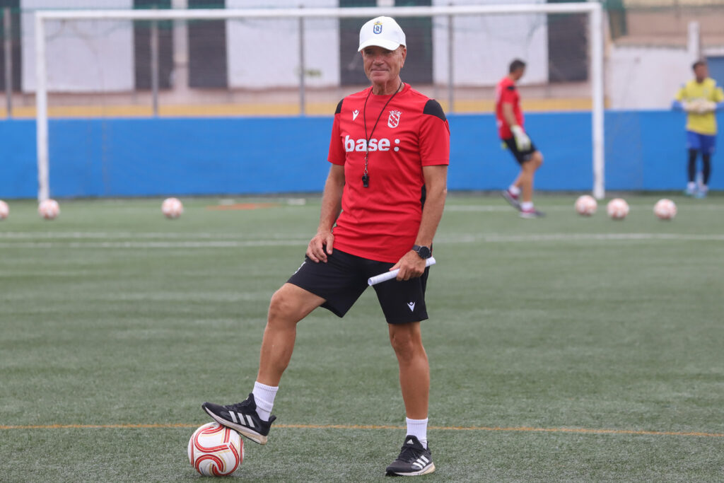 Soccer coach in a red shirt and white cap balances a ball with his foot during training on a green field, clipboard in hand.