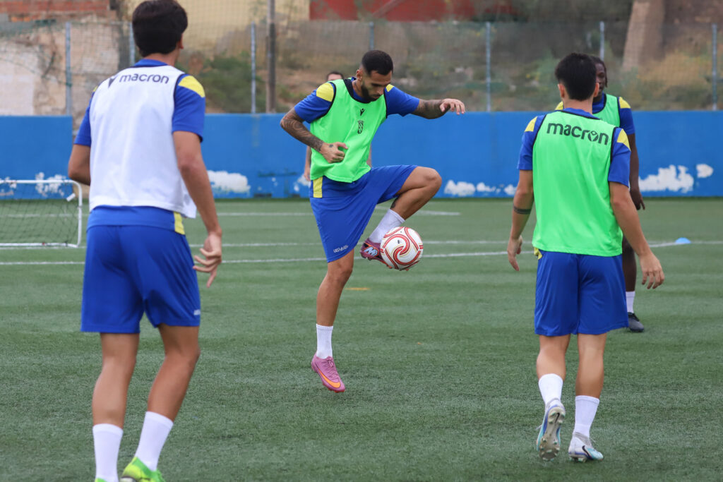 Soccer players in blue kit training on a turf field; central player in a green bib is kicking a ball mid-air while teammates watch.
