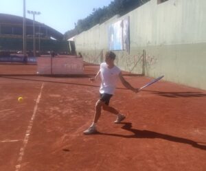 Tennis player in a white shirt and black shorts hitting a forehand on a red clay court, ball approaching on the left.