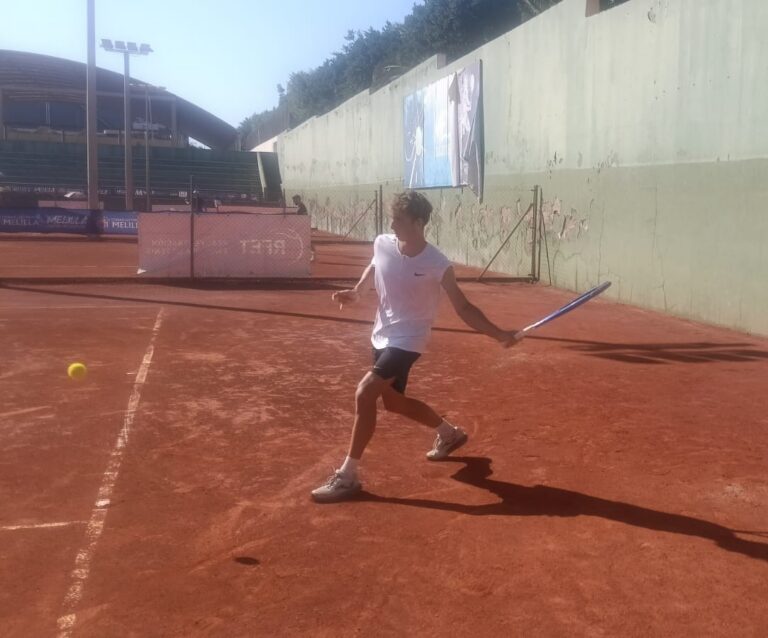 Tennis player in a white shirt and black shorts hitting a forehand on a red clay court, ball approaching on the left.