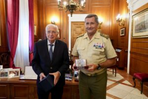 Two men in a formal office pose: a suited man holding a box while a uniformed officer presents a silver trophy on a wooden desk backdrop.