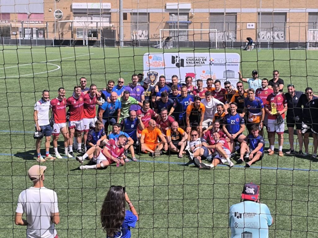 Group of soccer players posing for a team photo on an artificial turf field behind a fence, with a banner in the background.