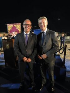 Two men in suits pose for a photo at a nighttime formal event, with a lectern and gold-embellished banner behind them behind them.