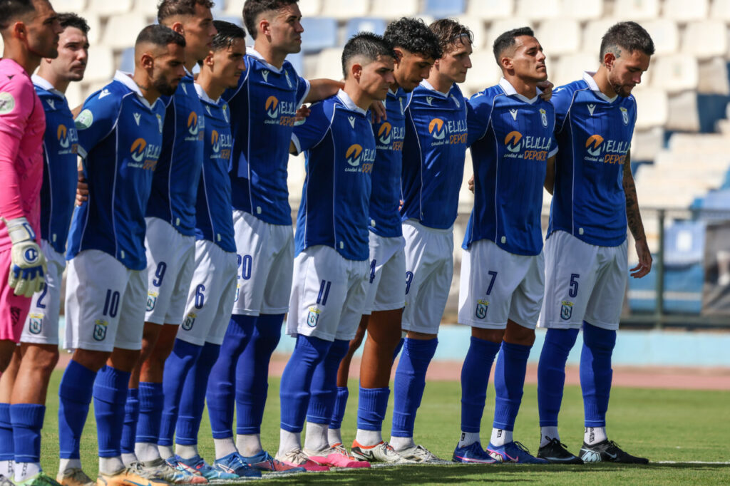 Soccer team lined up in blue uniforms on the field, arms around teammates before kickoff; goalkeeper in pink on the left.