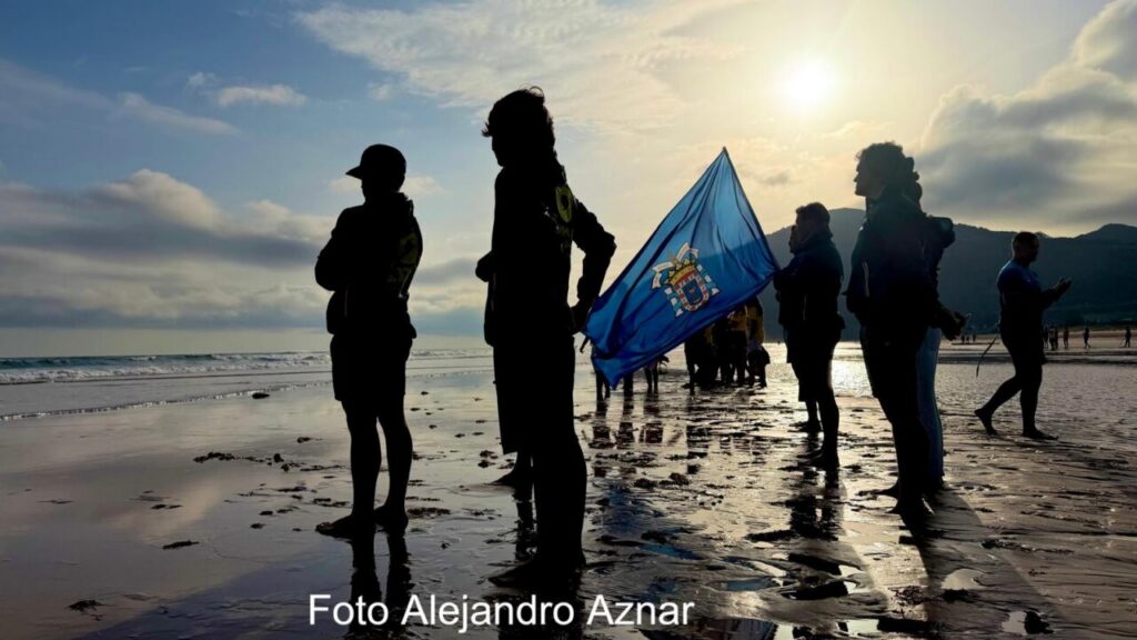 Silhouetted group on a wet beach at sunset, one person holding a blue flag with a crest.