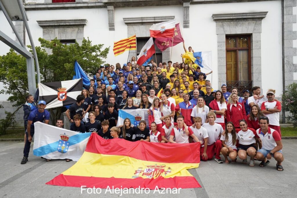 Large group of people posing with multiple national and regional flags in front of a stone building, suggesting a sports or cultural event venue delimited by trees and steps behind them.