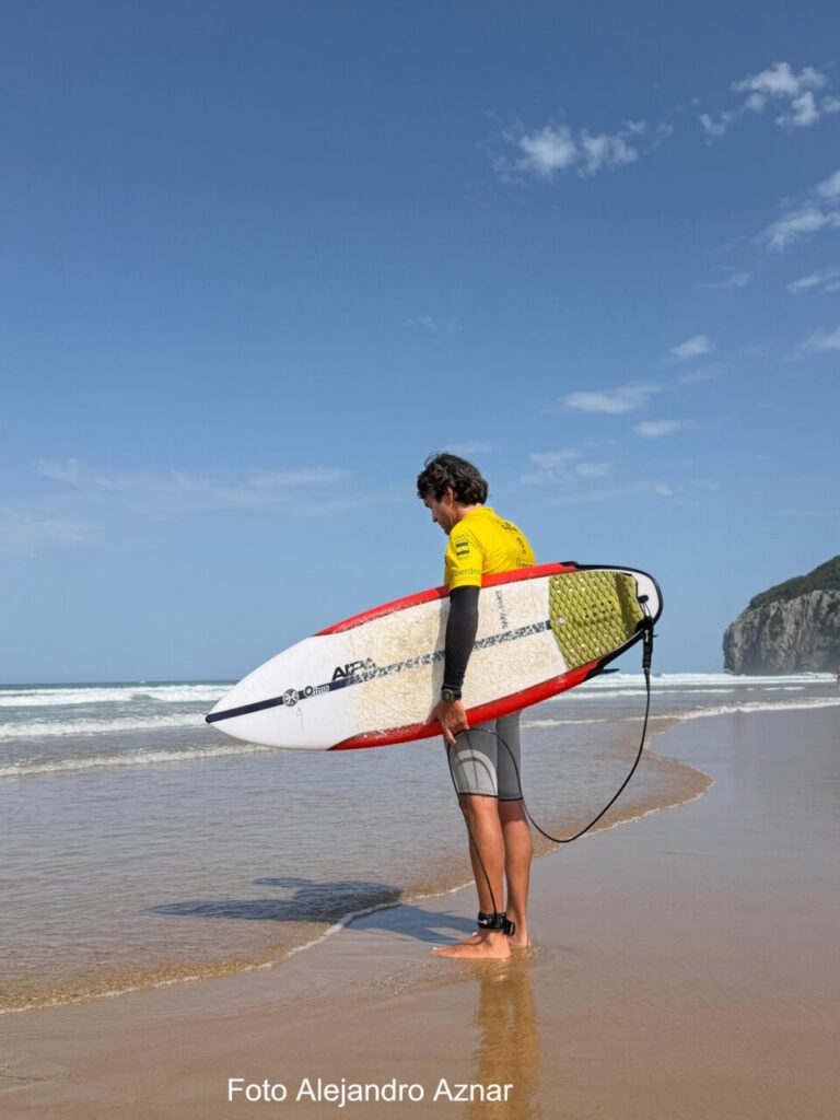 Surfer standing in shallow water on a sandy beach, holding a white and red surfboard with a leash, wearing a yellow rash guard and gray shorts, blue sky above.