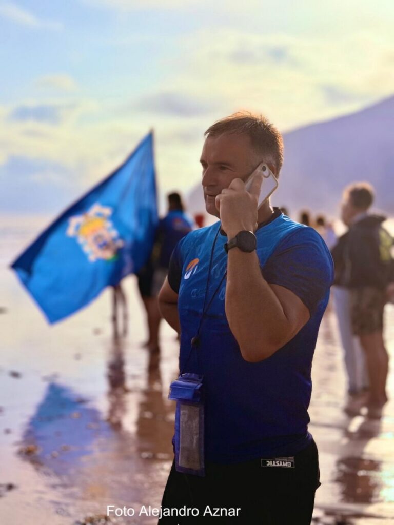 Man in a blue athletic shirt on a sunny beach talks on his cellphone, with a blue flag and other people in the background. He wears a lanyard and a water bottle holder hangs from his neck.