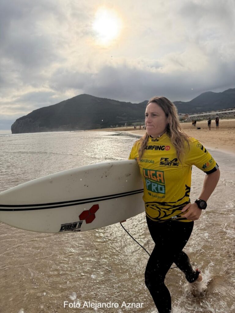 Woman in a yellow surfing jersey walks along the beach, carrying a white surfboard under her arm with mountains and a cloudy sky in the background.